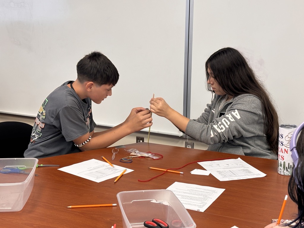 A young man and woman, seated at a table, work on a craft project using materials.