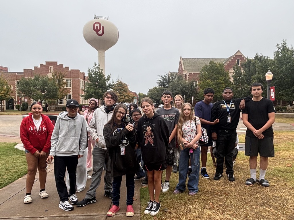 A group of people stand in front of a water tower, buildings, and trees on a lawn.