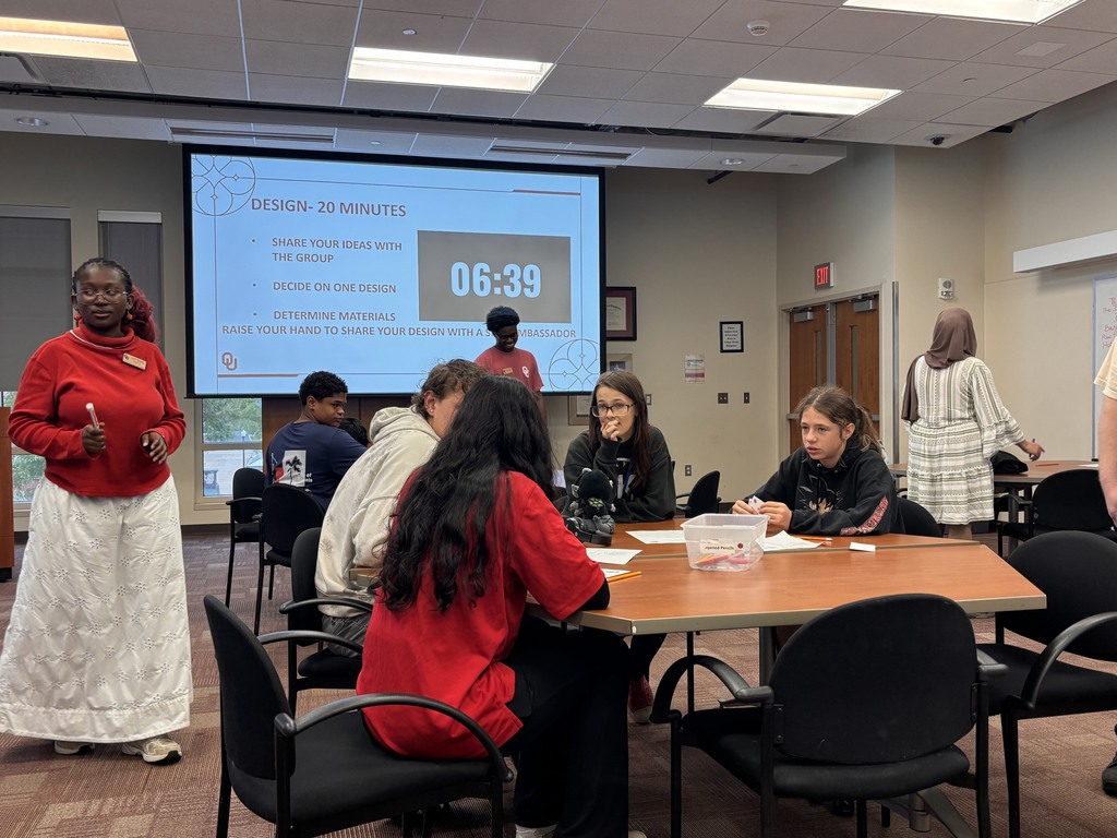 A classroom with a large screen displaying a timer reading 06:39. Several people sit around a table, and one stands by the table.