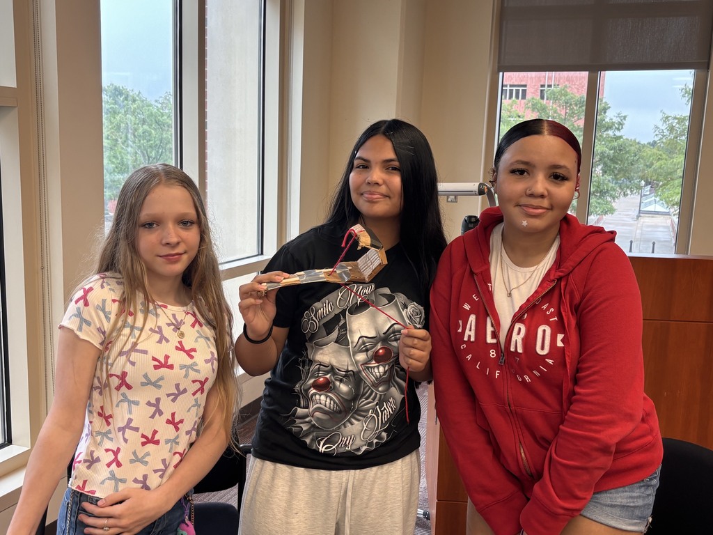 Three young women stand in a room with large windows, one holding a ribbon. They wear casual clothing.