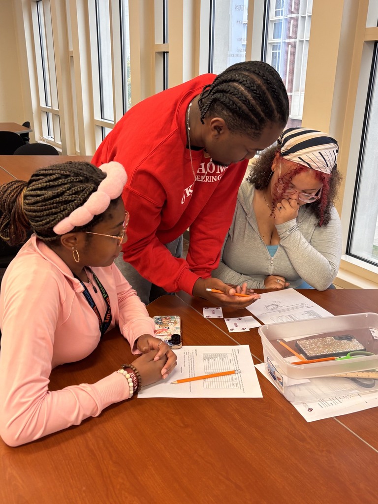 Three individuals collaborate on a project at a table with papers, pencils, and a plastic box.
