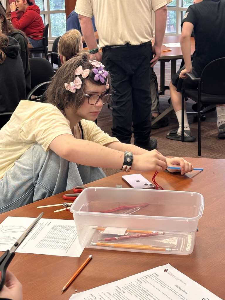 Woman with flowered hair and glasses sitting at a table with a box of pencils, papers, and scissors.