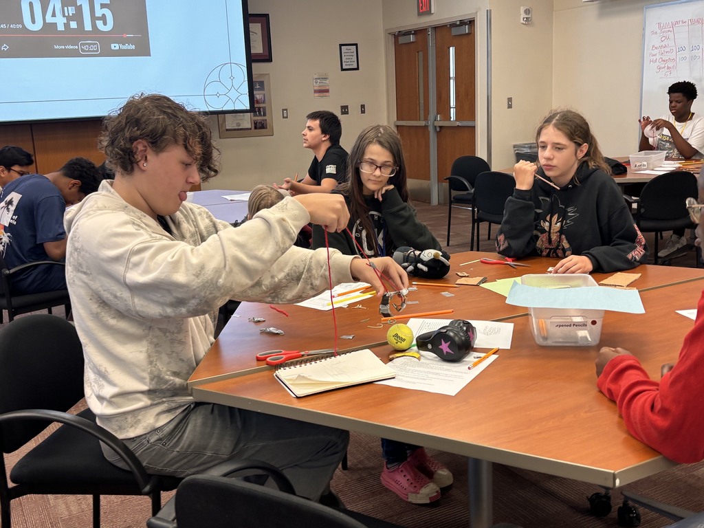 A group of students at a table in a classroom. Papers, scissors, and a ball are on the table.
