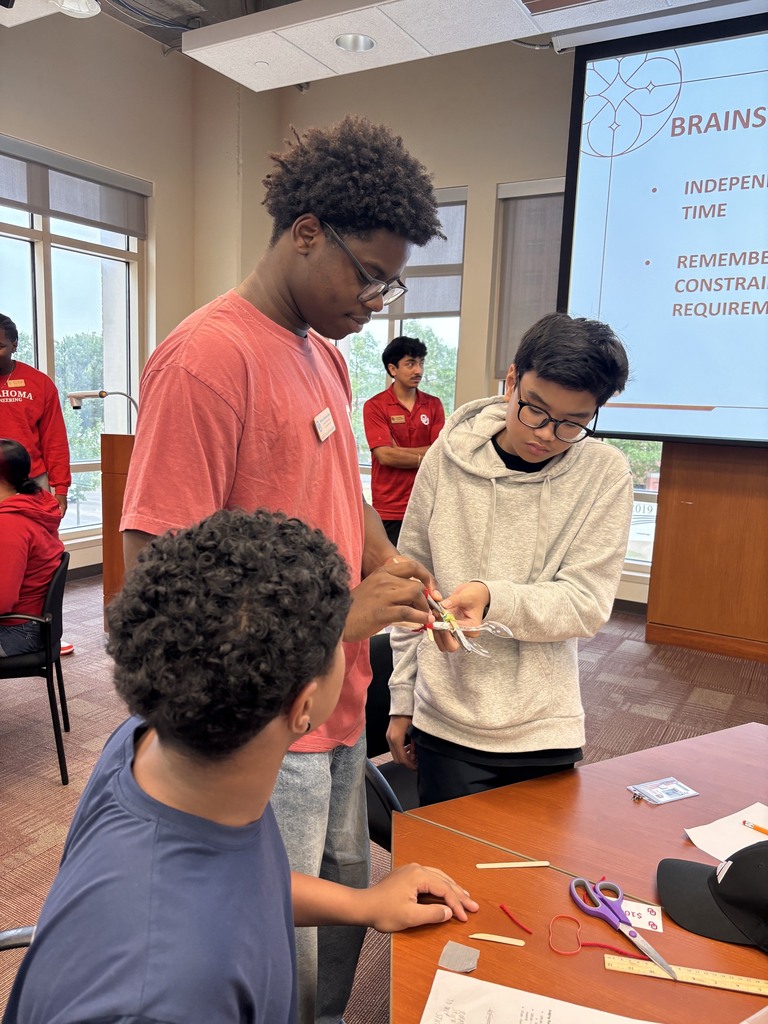 Three students focus on an activity at a table in a classroom. One holds a tool. Behind them, a screen displays "BRAINS" and "INDEPENDENT TIME."