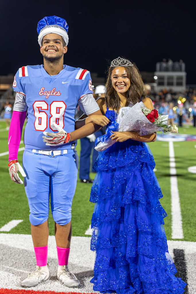 A football player in a blue uniform and a girl in a blue gown stand on a field, both smiling.