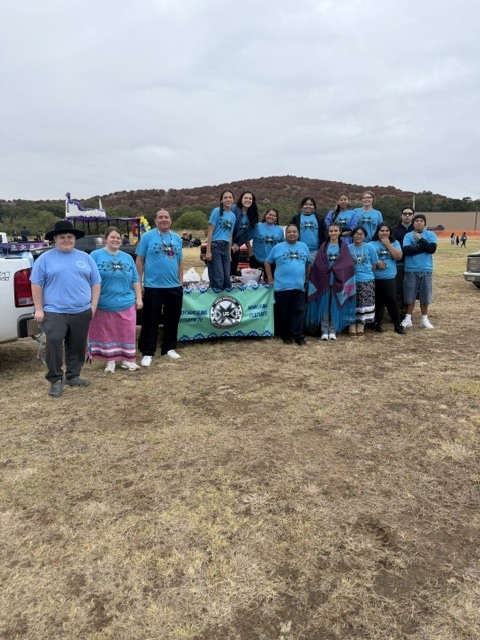 A group of people in blue shirts stands in a field. Some are seated on a table.