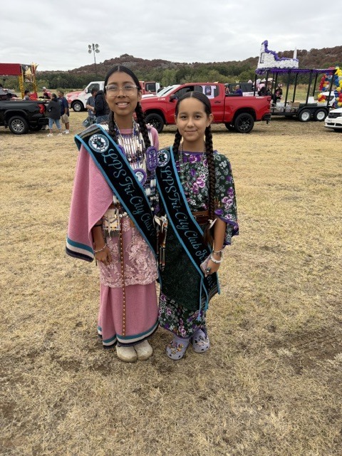 Two young girls stand in a field with traditional attire. One wears a pink cape, and the other has a floral dress. Vehicles and a trailer are in the background.