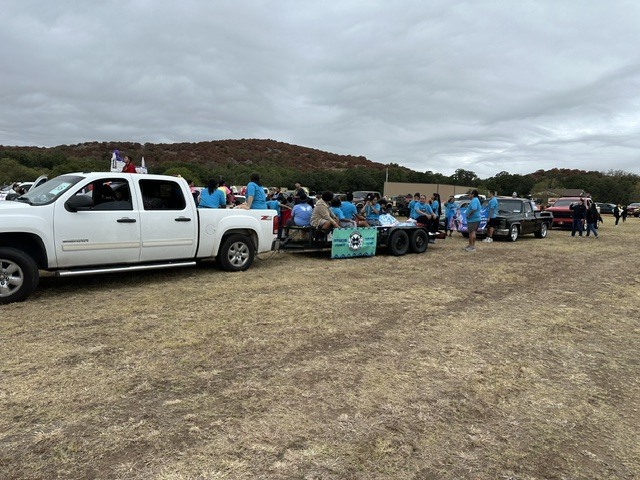 Vehicles parked in a field with a cloudy sky. People are standing around a pickup truck with a banner.
