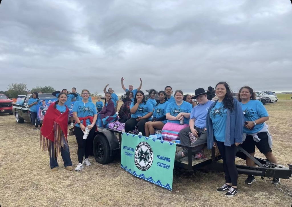 Group of people in matching blue shirts, some seated on a trailer, others standing, with a banner and cloudy sky in the background.