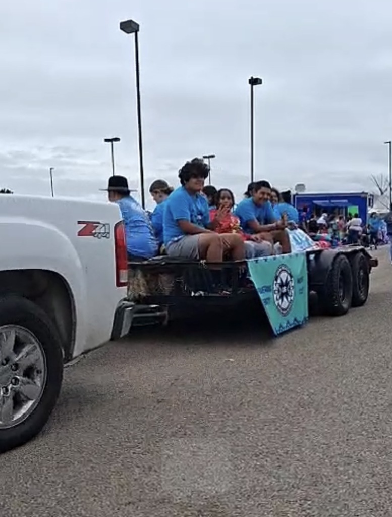 People in blue shirts sit on a trailer attached to a white pickup truck. A banner hangs behind them.