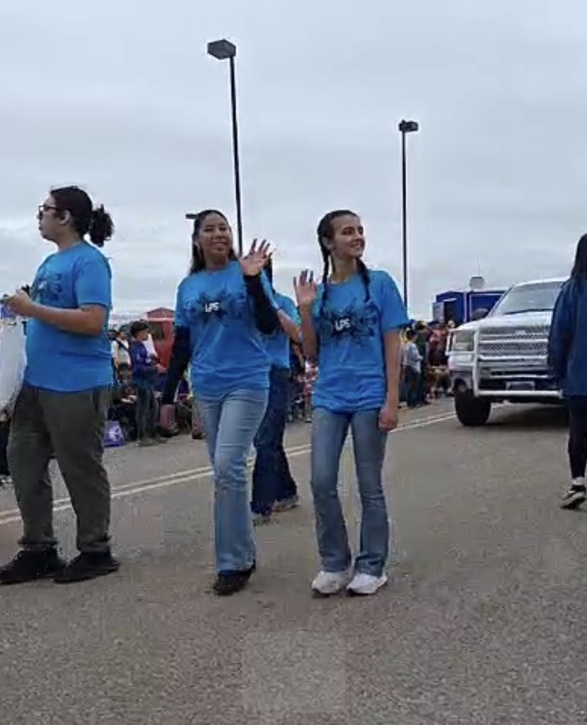 Three people in blue shirts and jeans walk along a road, waving. A pickup truck is behind them.