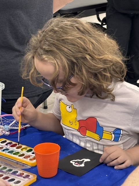 A child sits at a table with a blue tablecloth, wearing glasses and painting with a brush.