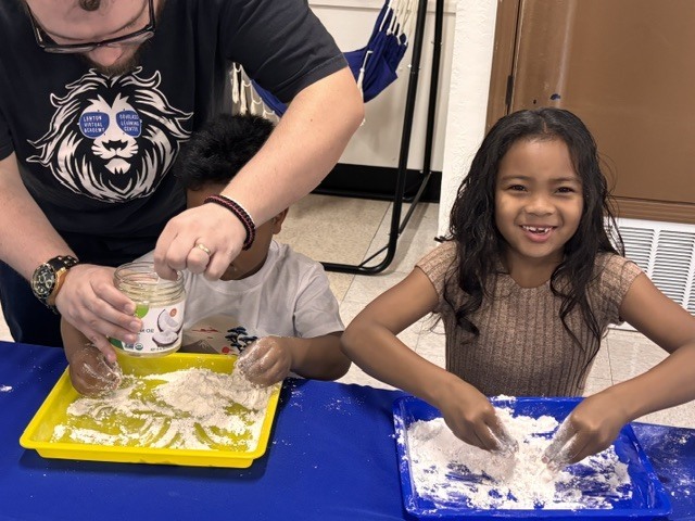 A man and two children are seated, engaging in an activity at a table with trays of sand.