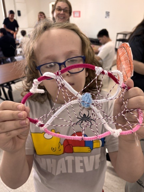 Child in a classroom holds up a pink and white web-like craft. Several people in background.