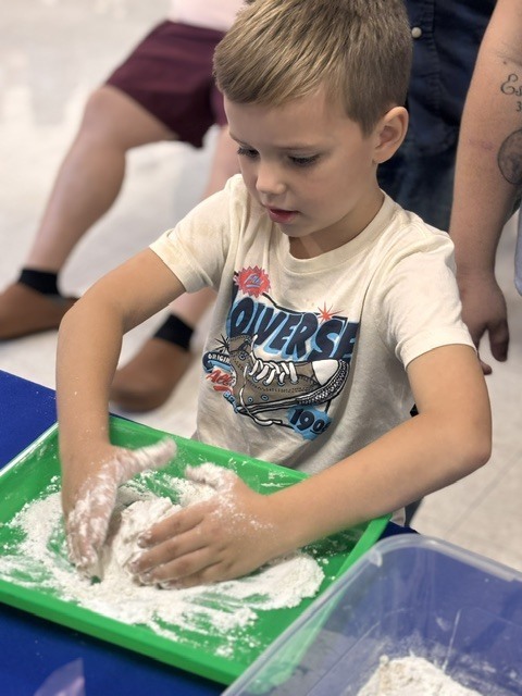 A young boy wearing a white shirt plays with white powder in a green tray. Behind him, two other people are seated, with one wearing brown boots.