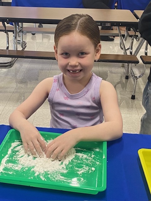 A girl with blonde hair wearing a purple shirt smiles and plays with green powder in a tray.