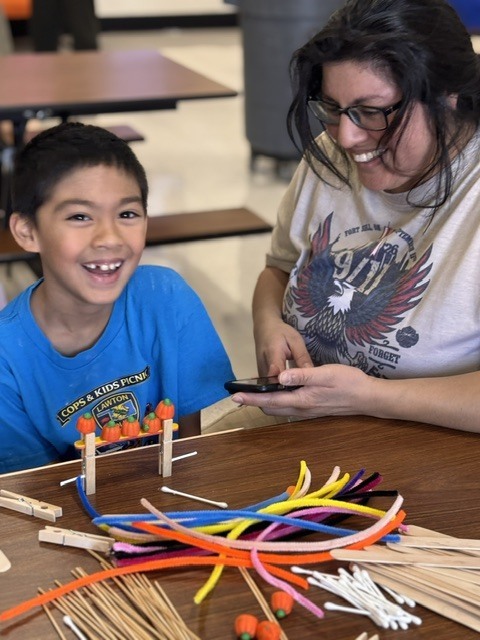 A woman and a boy work on a colorful, string project together at a table. The boy smiles.