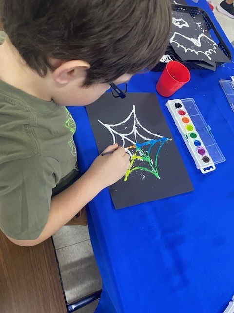 A child wearing a green shirt sits at a blue table, painting a spider web design on paper.