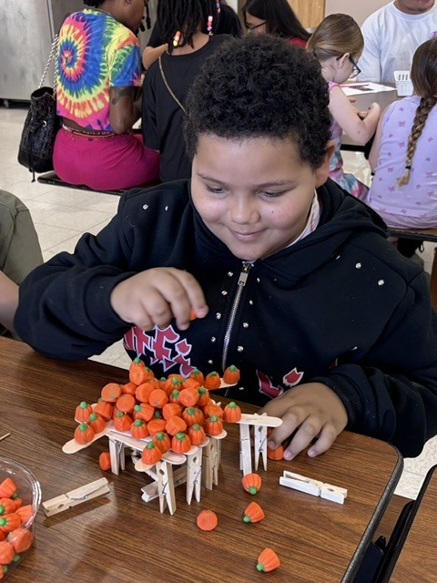 A child with curly hair sits at a table, placing orange candies on a small structure.