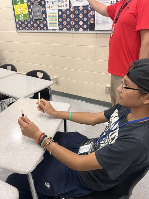 Two students in a classroom, one seated using a marker on a desk, the other standing with a marker.