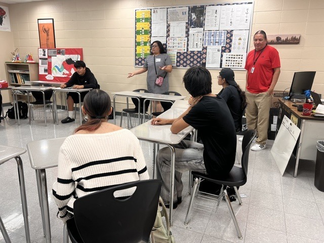 Students seated at desks, a woman at a desk speaks to a group. A teacher stands nearby, posters on walls, computers and books in background.