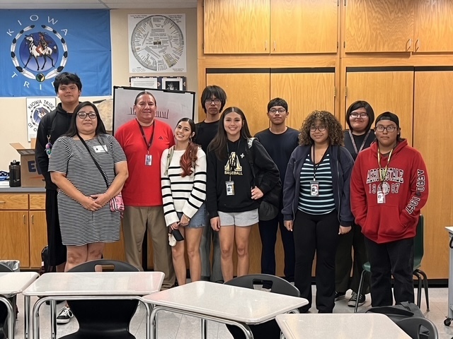 Students wearing IDs stand in a classroom with desks and cabinets, posing for a picture.