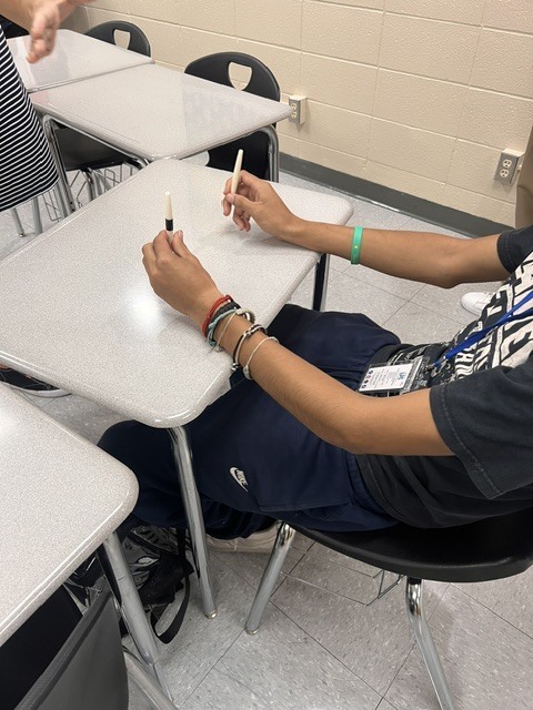 Student seated at desk holding two small plastic tubes with pencils, one in each hand.