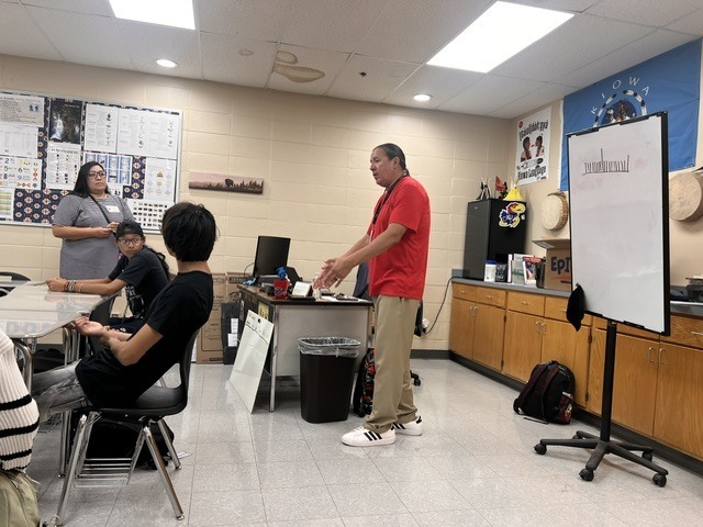 Adult standing in front of students in a classroom. A whiteboard is on a stand to the right.