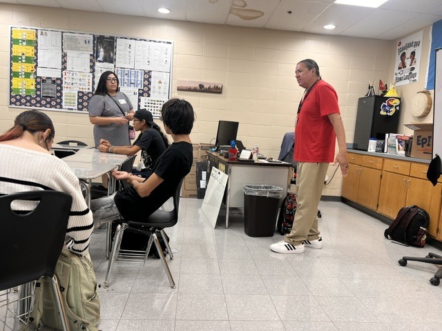 Several students seated at a table in a classroom while a teacher stands and speaks.