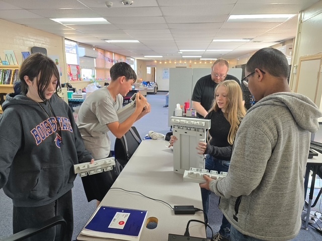 Several students in a classroom work around a table with a large electronic device.