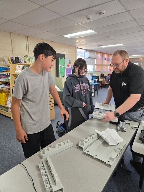 Three people examine pieces of equipment on a table; shelves with books and items in the background.