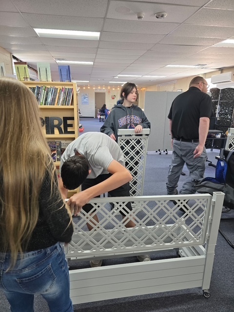 Students working on a white bed frame with a pattern in a room with bookcases and furniture.