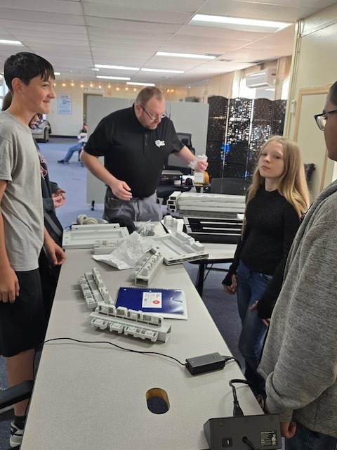 People stand around a table with a laptop, cables, and other objects on it. A person in glasses speaks.