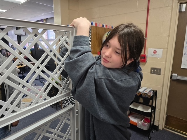 Girl holds up a white slat on a metal structure against a white wall.