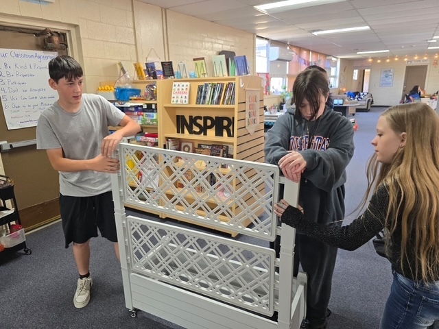 Three students stand around a white rack with a sign that says "Inspire" as a bookshelf sits behind them.