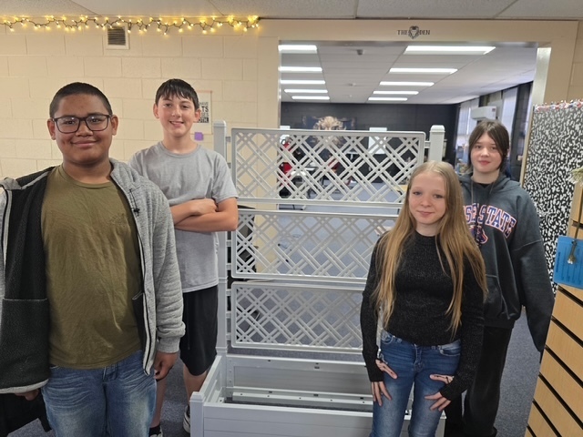 Four students standing near a white trellis in an indoor room with a wooden cabinet.