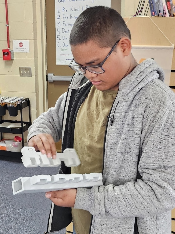 A boy in a gray hoodie examines a white, rectangular object with keys while standing in a classroom.