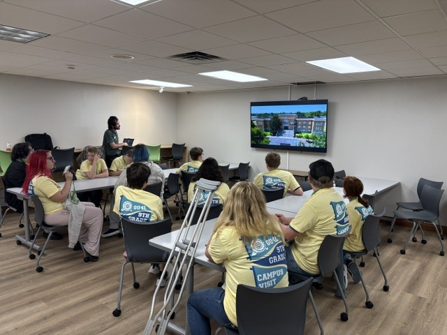 Group of people in yellow shirts sitting around tables, facing a screen on the wall, in a classroom.