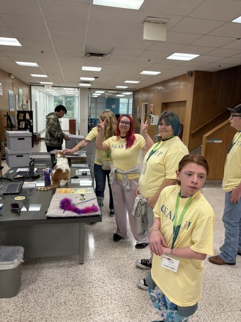 Six people in yellow shirts in a hall with a long desk. One person standing in front.