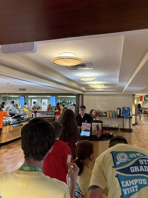 Several people wearing yellow shirts stand at a counter in a school cafeteria; people sit at tables behind them.