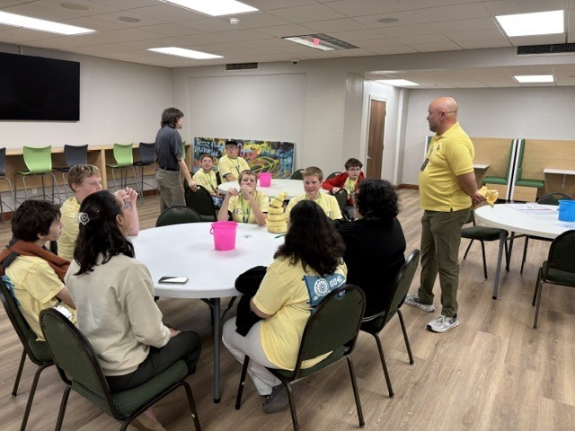 Group of students and teachers in a meeting room with tables, chairs, a TV, and a door.