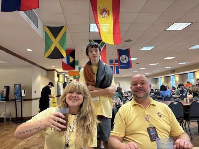 A room with various flags and people seated at a table. A woman holds a drink, and a man smiles at the camera.