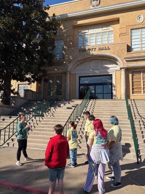 Several children and adults stand near a large building with a large tree and stairs.