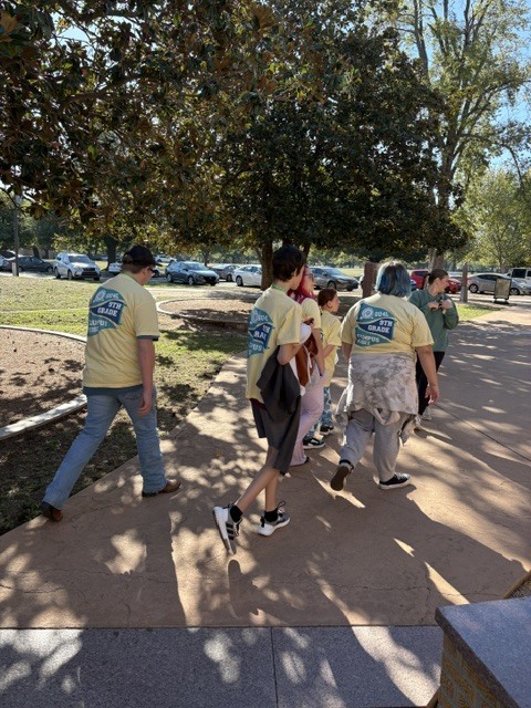 Five people in yellow shirts walking on a sidewalk with trees, green grass, and cars in the background.