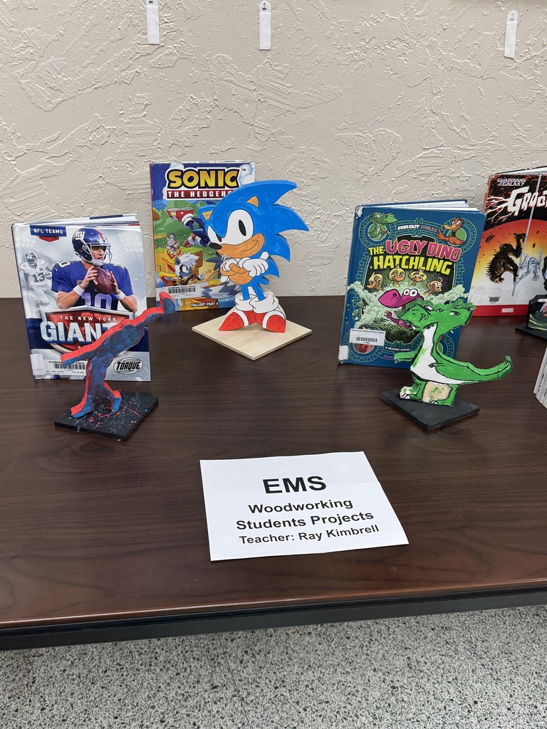 A table has books, figurines, and a sign. Books include Sonic, Giant, and others. Figurines include Sonic, a dinosaur, and a green creature.