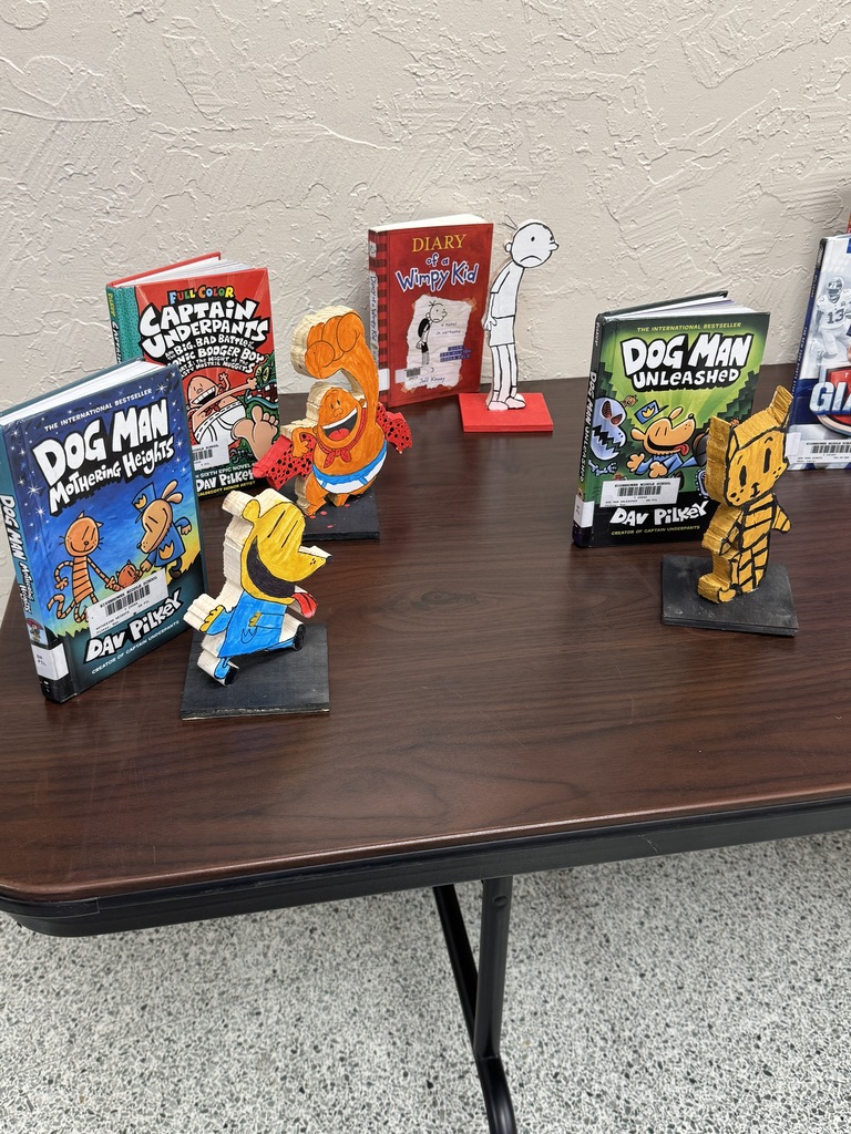 Books and figurines on a table. Titles include "Dog Man", "Captain Underpants", and "Big Nate".