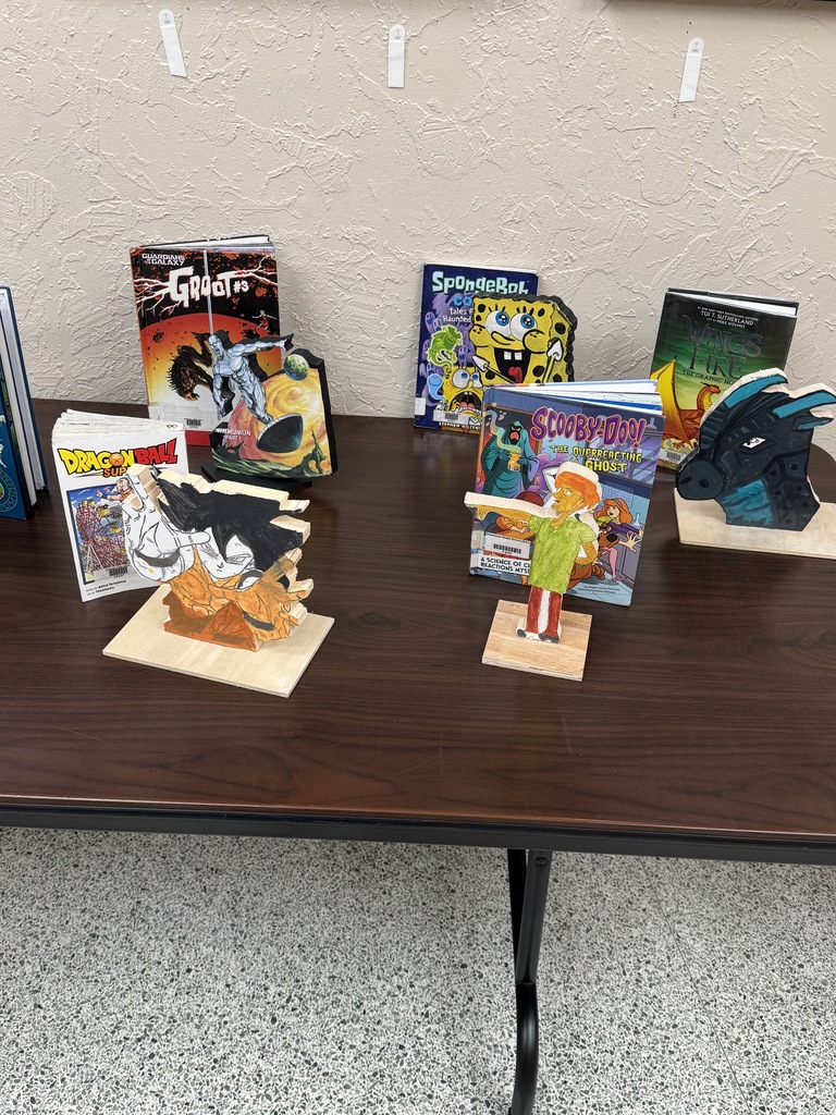 A brown table displays various books and crafted figurines, with a textured wall in the background.