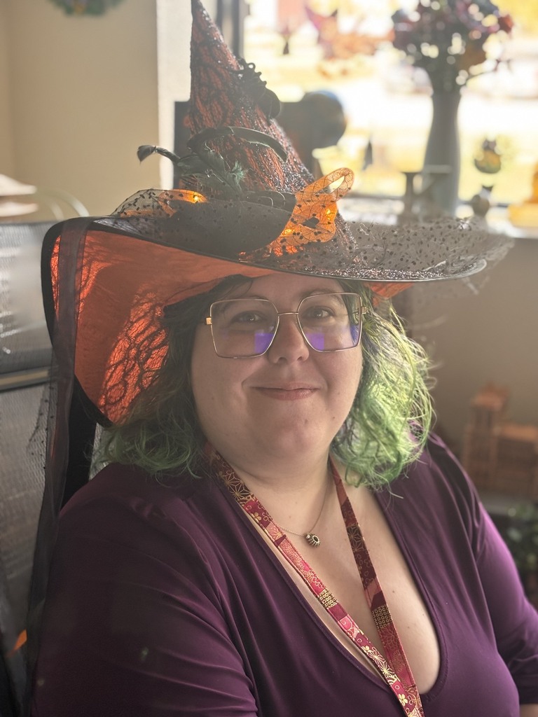 Woman wearing purple, smiling, sitting in chair, wearing glasses, witch hat with lace and orange butterfly.