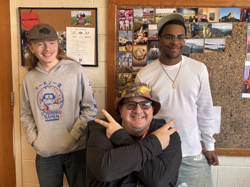 Three students standing near a bulletin board covered in photos. The middle one makes a peace sign.
