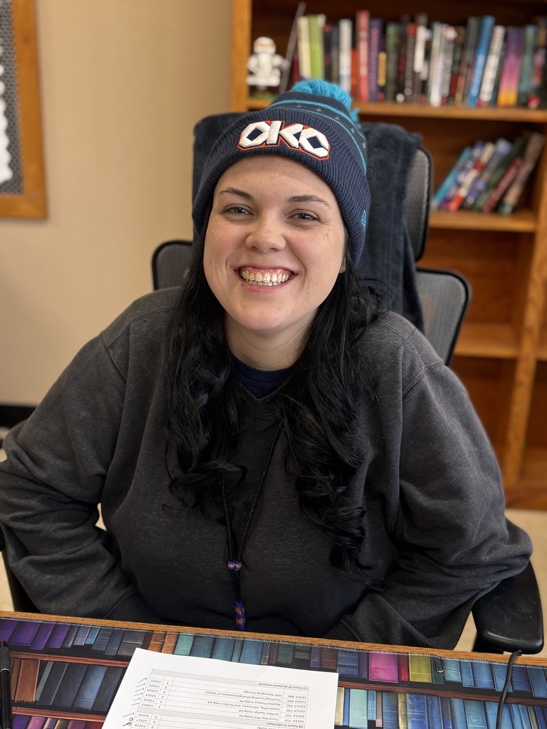 A woman smiles at the camera while sitting at a desk. Behind her, a bookshelf filled with books.